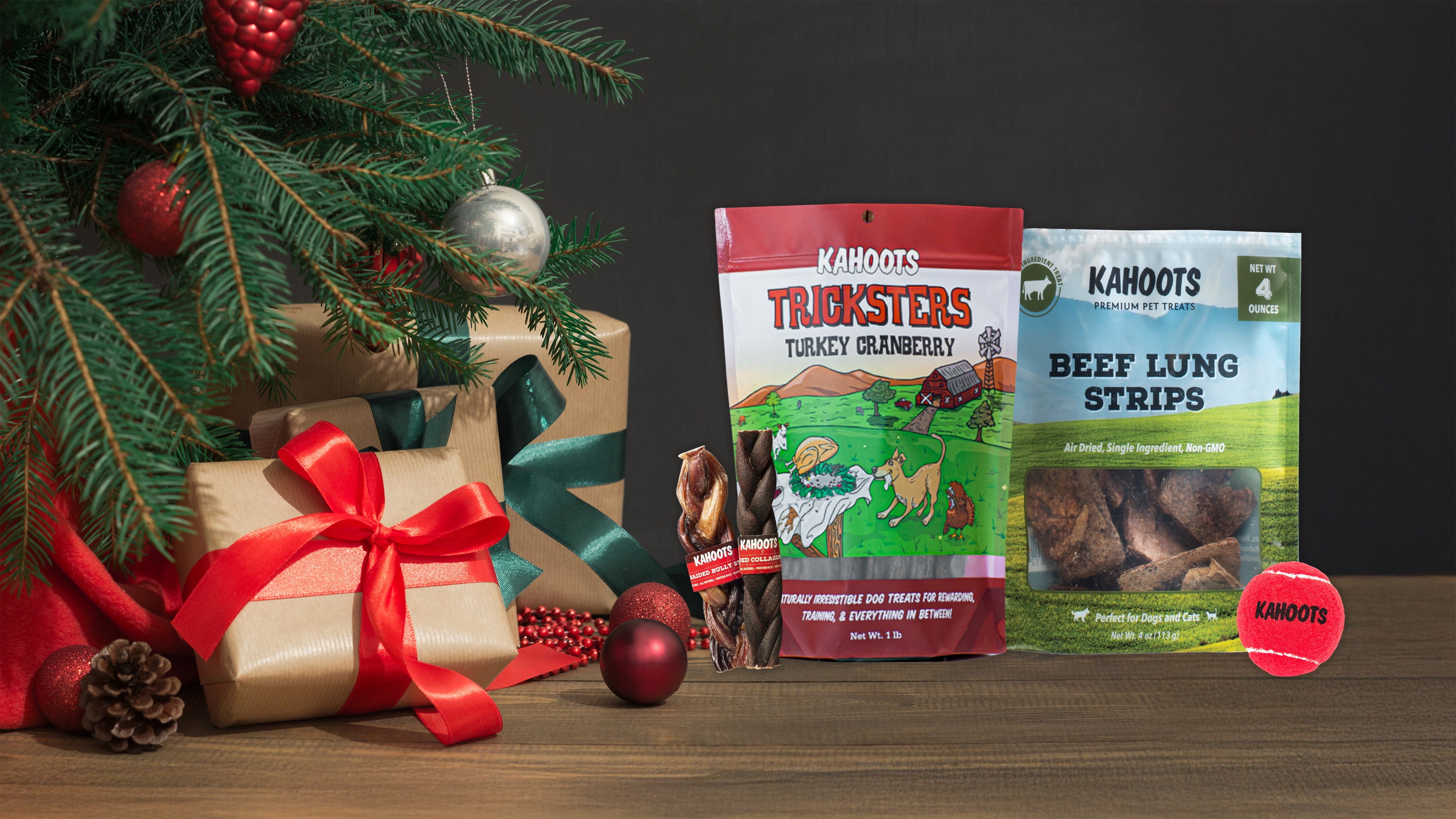 Two packages of dog TREATS next to a Christmas tree and wrapped gifts on a wooden surface.